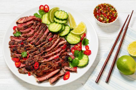 crying tiger, grilled and thinly sliced ribeye steak with cucumber and lime on plate on white wooden table with spicy dipping sauce, thai cuisine