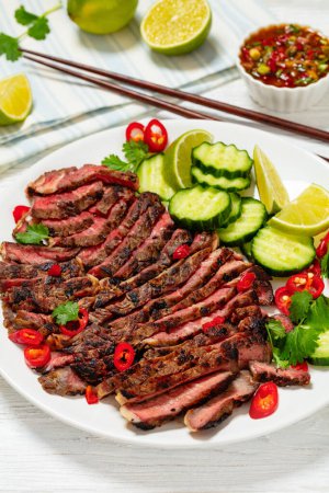 crying tiger, grilled and thinly sliced ribeye steak with cucumber and lime on plate on white wooden table with spicy dipping sauce, thai cuisine, vertical view from above