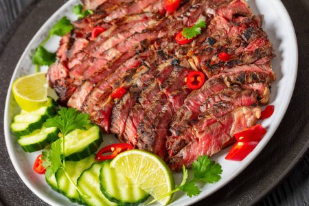 crying tiger, grilled and thinly sliced ribeye steak with cucumber and lime on plate on black wooden table, thai cuisine, close-up, dutch angle view