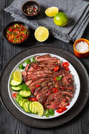 crying tiger, grilled and thinly sliced ribeye steak with cucumber and lime on plate on black wooden table with spicy dipping sauce, thai cuisine, vertical view from above