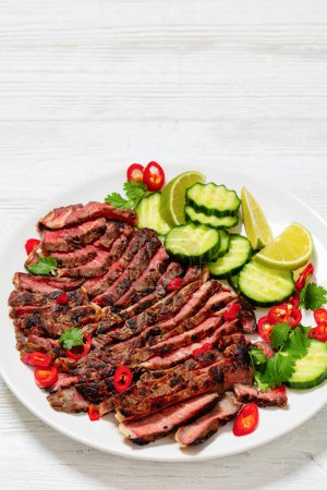 crying tiger, grilled and thinly sliced ribeye steak with cucumber and lime on plate on white wooden table with spicy dipping sauce, thai cuisine, vertical view, free space