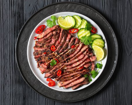 crying tiger, grilled and thinly sliced ribeye steak with cucumber and lime on plate on black wooden table with spicy dipping sauce, thai cuisine, horizontal view from above, flat lay, close-up