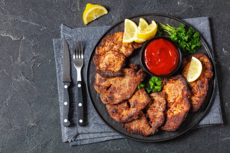fried juicy pork steaks with ketchup, lemon slices and parsley on black plate on concrete table with cutlery, horizontal view from above, flat lay, free space