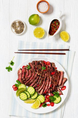 crying tiger, grilled and thinly sliced ribeye steak with cucumber and lime on plate on white wooden table with spicy dipping sauce, thai cuisine, vertical view from above, flat lay