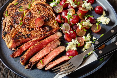 gebratenes Rindersteak serviert mit Salat aus roten Trauben, zerbröseltem Blauschimmelkäse und Schnittlauch auf Teller mit Besteck auf dunklem Holztisch, Nahaufnahme,