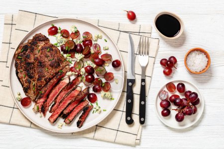 gebratenes Rindersteak mit rotem Traubensalat, zerbröseltem Blauschimmelkäse und Schnittlauch auf Teller auf weißem Holztisch mit Besteck, horizontale Sicht von oben, flache Lage