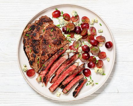 Roastbeef-Steak mit Salat aus roter Traube, zerbröseltem Blauschimmelkäse und Schnittlauch auf Teller auf weißem Holztisch, horizontale Sicht von oben, flache Lage, Nahaufnahme