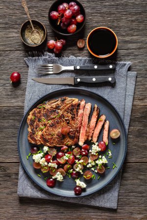 gebratenes Rindersteak serviert mit Salat aus roter Traube, zerbröseltem Blauschimmelkäse und Schnittlauch auf Teller auf dunklem Holztisch mit Besteck, vertikaler Blick von oben, flache Lage