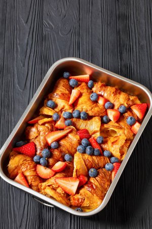 strawberry blueberry croissant casserole in baking dish on black wooden table, vertical view from above, free space