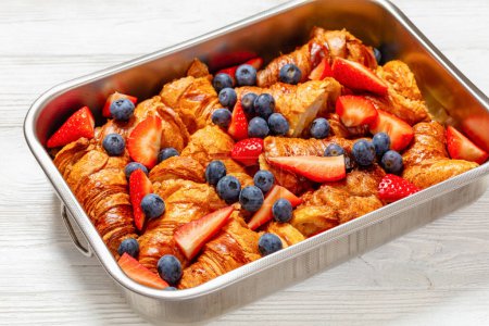 strawberry blueberry croissant casserole in baking dish on white wooden table, horizontal view from above, close-up