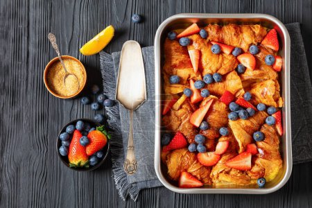 Erdbeer-Blaubeer-Croissant-Auflauf in Auflaufform auf schwarzem Holztisch mit Zutaten und Kuchenschaufel, horizontale Sicht von oben, flache Lage