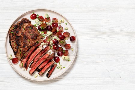 Roastbeef-Steak mit Salat aus roter Traube, zerbröseltem Blauschimmelkäse und Schnittlauch auf Teller auf weißem Holztisch, horizontale Sicht von oben, flache Lage, Nahaufnahme