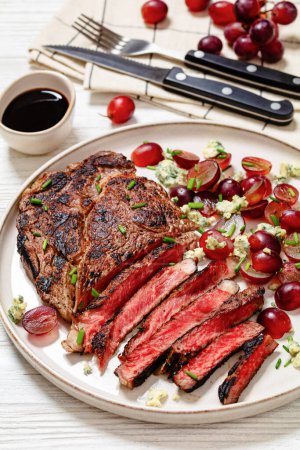 gebratenes Rindersteak mit rotem Traubensalat, zerbröseltem Blauschimmelkäse und Schnittlauch auf Teller auf weißem Holztisch mit Besteck, vertikale Sicht von oben, Nahaufnahme