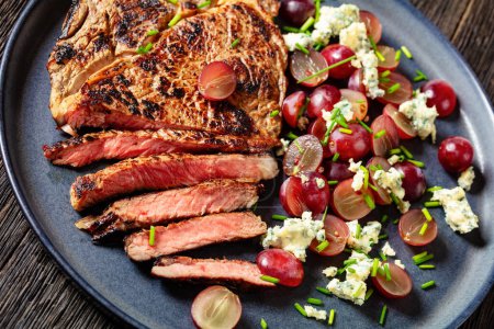 gebratenes Rindersteak serviert mit Salat aus roten Trauben, zerbröseltem Blauschimmelkäse und Schnittlauch auf Teller auf dunklem Holztisch, Nahaufnahme,
