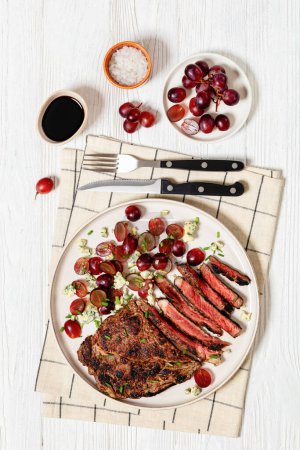 gebratenes Rindersteak serviert mit Salat aus roten Trauben, zerbröseltem Blauschimmelkäse und Schnittlauch auf Teller auf weißem Holztisch mit Besteck, vertikaler Blick von oben