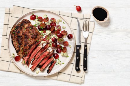 roast beef steak served with salad of red grape, crumbled blue mold cheese and chives on plate on white wooden table with cutlery, horizontal view from above, flat lay, free space