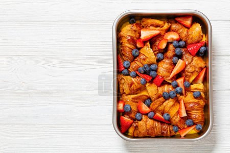 strawberry blueberry croissant casserole in baking dish on white wooden table, horizontal view from above, flat lay, free space