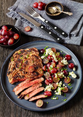 gebratenes Rindersteak serviert mit Salat aus roter Traube, zerbröseltem Blauschimmelkäse und Schnittlauch auf Teller auf dunklem Holztisch mit Besteck, vertikaler Blick von oben