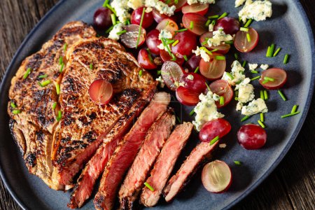 gebratenes Rindfleisch-Entrecote serviert mit Salat aus roter Traube, zerbröseltem Blauschimmelkäse und Schnittlauch auf Teller auf dunklem Holztisch, holländischer Blickwinkel, Nahaufnahme,
