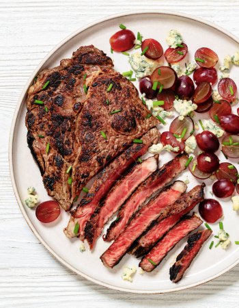 Roastbeef Steak serviert mit Salat aus roten Trauben, zerbröseltem Blauschimmelkäse und Schnittlauch auf Teller auf weißem Holztisch, Großaufnahme, vertikale Ansicht