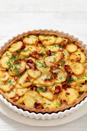 Irish potato pie of crispy crust layered with potatoes, bacon and onion in baking dish on white wooden table, vertical view from above, close-up, free space