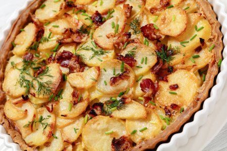 close-up of Irish potato pie of crispy crust layered with potatoes, bacon and onion in baking dish on white wooden table, dutch angle view