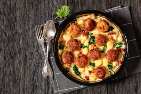 tuscan chicken meatballs with gnocchi in creamy sauce with sun-dried tomatoes and spinach leaves in baking dish on dark wooden table with fork and spoon, horizontal view from above, flat lay