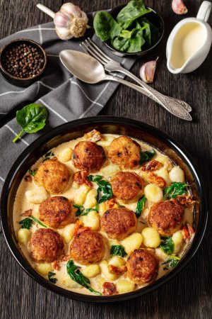 tuscan chicken meatballs with gnocchi in creamy sauce with sun-dried tomatoes and spinach leaves in baking dish on dark wooden table with ingredients, vertical view