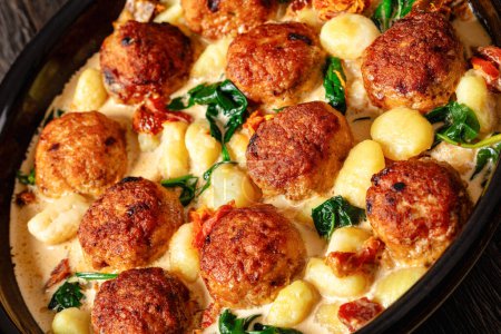 close-up of tuscan chicken meatballs with gnocchi in creamy sauce with sun-dried tomatoes and spinach leaves in baking dish