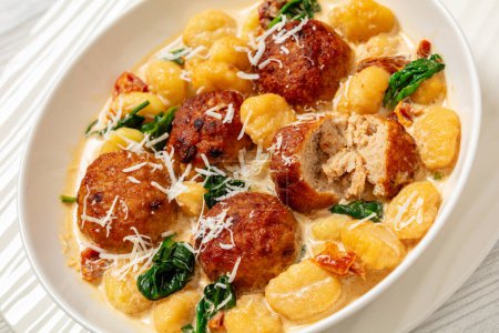 close-up of tuscan chicken meatballs with gnocchi in creamy sauce with sun-dried tomatoes and spinach leaves in white bowl on white wooden table, dutch angle view