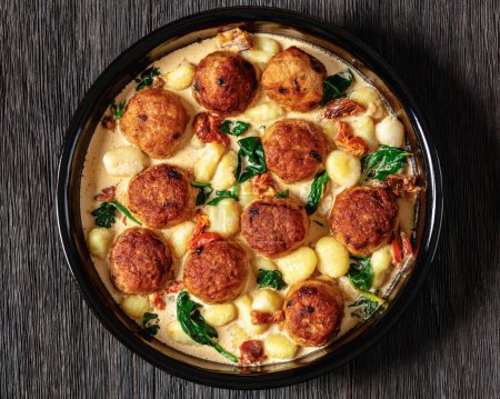 tuscan chicken meatballs with gnocchi in creamy sauce with sun-dried tomatoes and spinach leaves in baking dish on dark wooden table, horizontal view from above, flat lay, close-up
