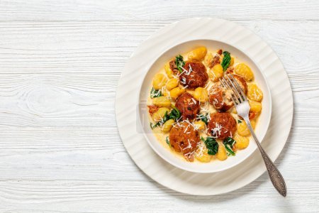 tuscan chicken meatballs with gnocchi in creamy sauce with sun-dried tomatoes and spinach leaves in white bowl on white wooden table with fork, horizontal view from above, flat lay, free space