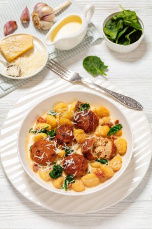 tuscan chicken meatballs with gnocchi in creamy sauce with sun-dried tomatoes and spinach leaves in white bowl on white wooden table with fork and ingredients, vertical view from above