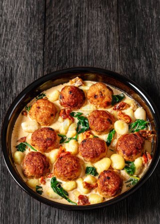 tuscan chicken meatballs with gnocchi in creamy sauce with sun-dried tomatoes and spinach leaves in baking dish on dark wooden table, vertical view, free space