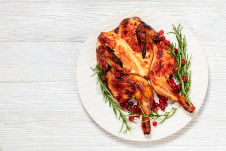 butterflied or spatchcock roast whole chicken on white plate with rosemary and cranberry on wooden table, horizontal view from above, flat lay, free space