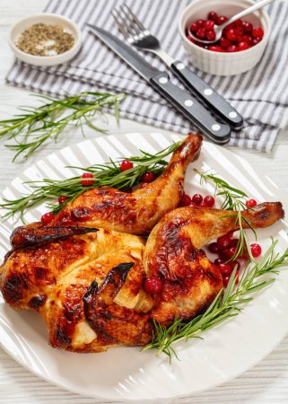 butterflied or spatchcock roast whole chicken on white plate with rosemary and cranberry on wooden table with cutlery, vertical view from above, close-up