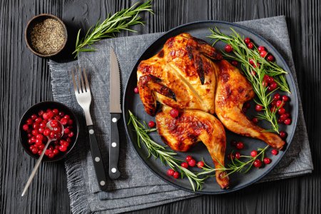 butterflied or spatchcock roast whole chicken on dark grey plate with rosemary and cranberry on black wooden table with cutlery and ingredients, flat lay