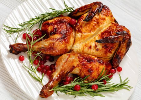butterflied or spatchcock roast whole chicken on white plate with rosemary and cranberry on wooden table, close-up