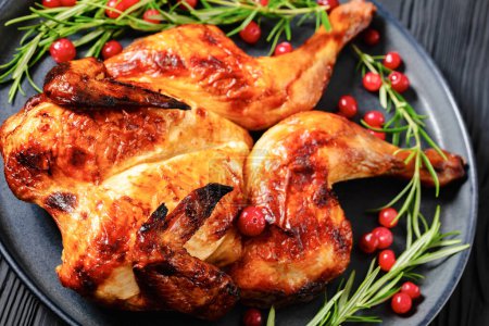 butterflied or spatchcock roast whole chicken on dark grey plate with rosemary and cranberry on black wooden table, close-up