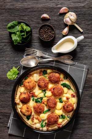 tuscan chicken meatballs with gnocchi in creamy sauce with sun-dried tomatoes and spinach leaves in baking dish on dark wooden table with ingredients, vertical view from above