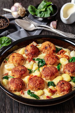 tuscan chicken meatballs with gnocchi in creamy sauce with sun-dried tomatoes and spinach leaves in baking dish on dark wooden table with ingredients, vertical view, close-up