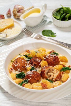 tuscan chicken meatballs with gnocchi in creamy sauce with sun-dried tomatoes and spinach leaves in white bowl on white wooden table with fork, horizontal view from above,close-up