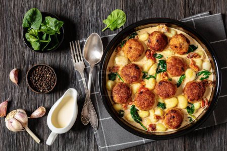 tuscan chicken meatballs with gnocchi in creamy sauce with sun-dried tomatoes and spinach leaves in baking dish on dark wooden table with ingredients, horizontal view from above, flat lay