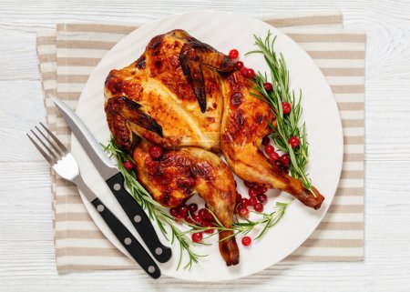 butterflied or spatchcock roast whole chicken on white plate with rosemary and cranberry on wooden rustic table with cutlery, horizontal view from above, flat lay, close-up