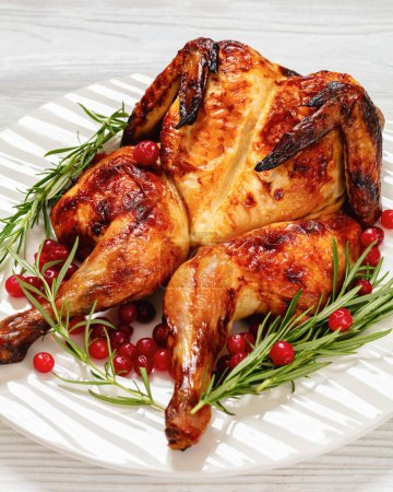butterflied or spatchcock roast whole chicken on white plate with rosemary and cranberry on wooden table, vertical view from above, close-up