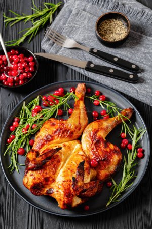 butterflied or spatchcock roast whole chicken on dark grey plate with rosemary and cranberry on black wooden table with cutlery, vertical view from above, close-up