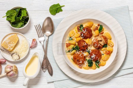 tuscan chicken meatballs with gnocchi in creamy sauce with sun-dried tomatoes and spinach leaves in white bowl on white wooden table with ingredients, horizontal view from above, flat lay