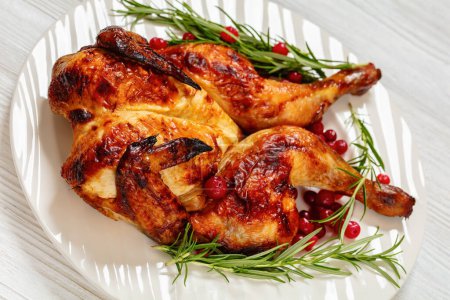 close-up of butterflied or spatchcock roast whole chicken on white plate with rosemary and cranberry on wooden table