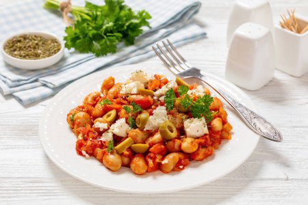 gigantes plaki, greek giant baked beans in a chunky tomato sauce sprinkled with feta cheese, olives, and fresh parsley on plate with fork on white wooden table, landscape view, close-up