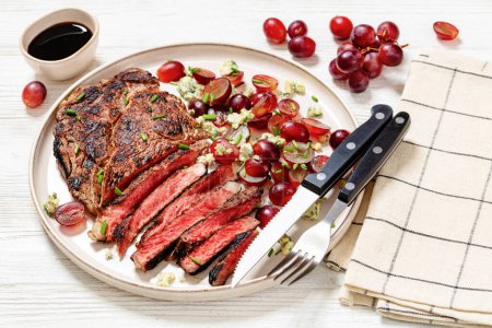 pan fried beef steak served with salad of red grape, crumbled blue mold cheese and chives on plate with cutlery on white wooden table, close-up